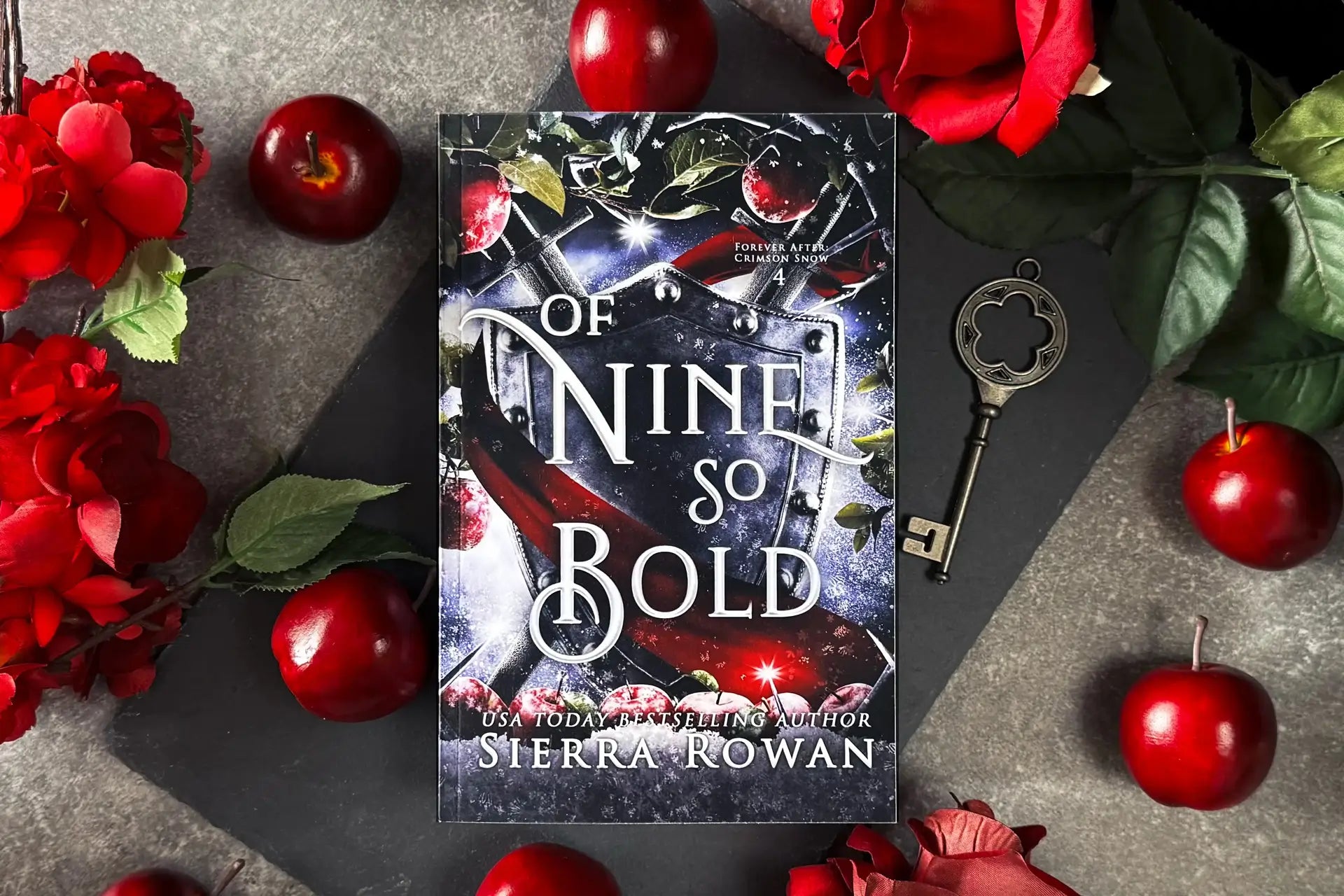 Of Nine So Bold by Sierra Rowan, paperback edition, on a slate board with apples and red flowers surrounding the book