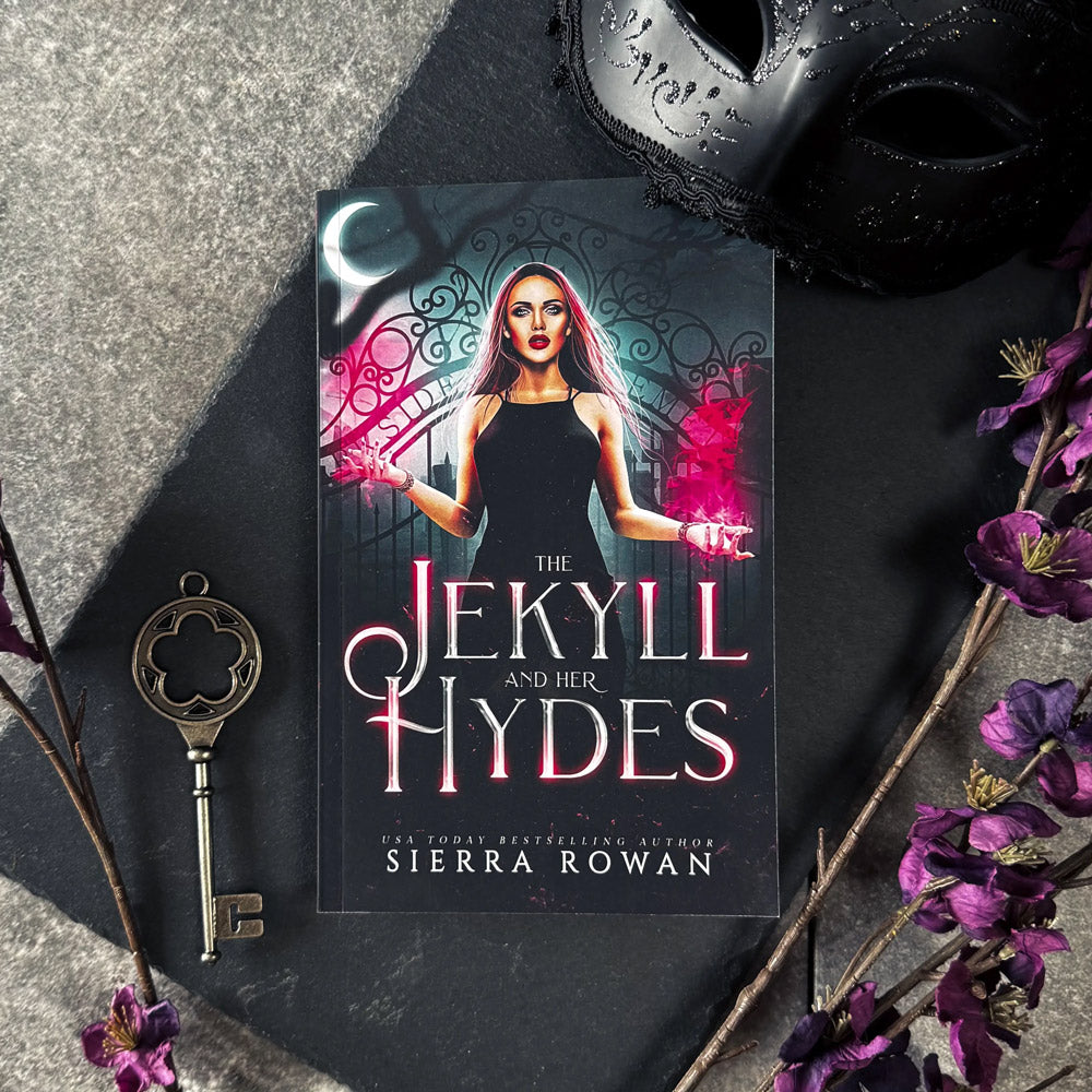 Paperback edition of The Jekyll and Her Hydes by USA Today Bestselling Author Sierra Rowan lays on a black slate board with a carnival mask above it and a gold key beside purple flowers at its side.