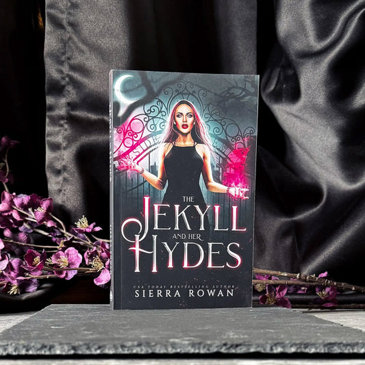 Paperback edition of The Jekyll and Her Hydes by Sierra Rowan stands upright against a black curtain with purple flowers behind the book and a slate board beneath the book.